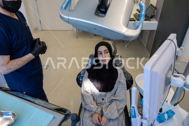 تقديم الاستشارات والنصائح الطبية، العمل في مجال جراحة الفم والفكين، صورة من الأعلى لمريضة مستلقية على كرسي العلاج في العيادة السنية وطبيب أسنان عربي خليجي سعودي يرتدي اليونيفورم والقفازات يقوم بشرح الحالة لها، استخدام الأدوات المعقمة والتقنيات والأجهزة الحديثة