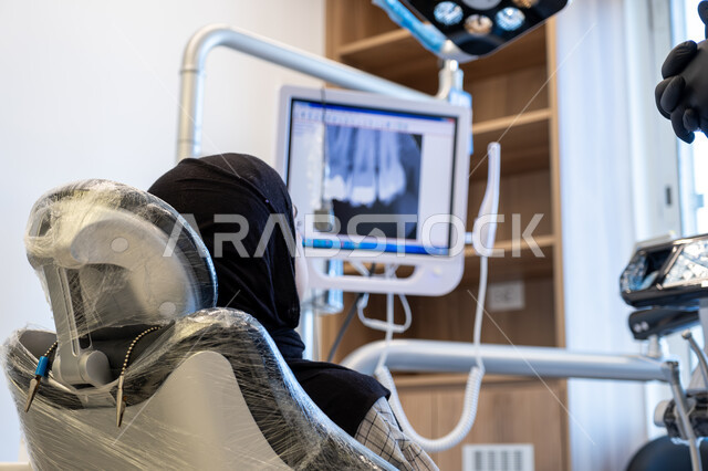 أهمية زيارة طبيب الأسنان بشكل دوري، تقديم الاستشارات والنصائح الطبية، العمل في مجال جراحة الفم والفكين، صورة من الخلف لمريضة عربية خليجية سعودية محجبة مستلقية على كرسي العلاج في العيادة السنية تنتظر بدء العلاج، الأدوات المعقمة والتقنيات والأجهزة الحديثة