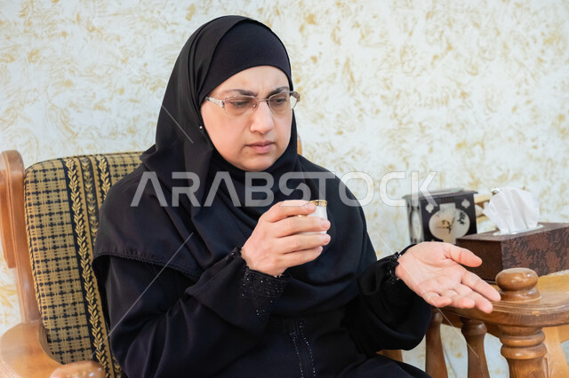 الإشارة باليدين والتحدث مع شخص ما، تناول المشروبات الساخنة اللذيذة، صورة مقربة لإمرأة عربية خليجية سعودية ترتدي الحجاب والعباءة التقليدية وتضع النظارة الطبية تجلس في غرفة المعيشة وتمسك بيدها فنجان من القهوة الفاخرة، إكرام الضيف وحسن استقباله