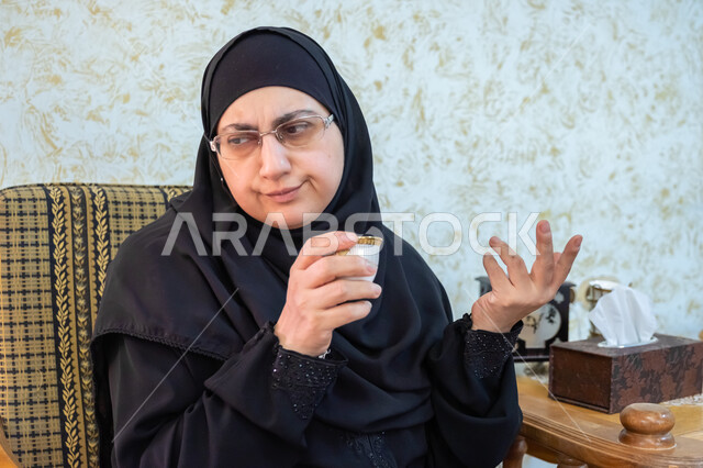 الإشارة باليدين والتحدث مع شخص ما، تناول المشروبات الساخنة اللذيذة، صورة مقربة لإمرأة عربية خليجية سعودية ترتدي الحجاب والعباءة التقليدية وتضع النظارة الطبية تجلس في غرفة المعيشة وتمسك بيدها فنجان من القهوة الفاخرة، إكرام الضيف وحسن استقباله
