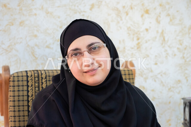 النظر للكاميرا بإيماءات البشاشة والسرور والرضا، صورة مقربة لإمرأة عربية خليجية سعودية ترتدي الحجاب والعباءة التقليدية وتضع النظارة الطبية تجلس على أريكة خشبية، الجلوس في غرفة المعيشة والحصول على بعض الراحة