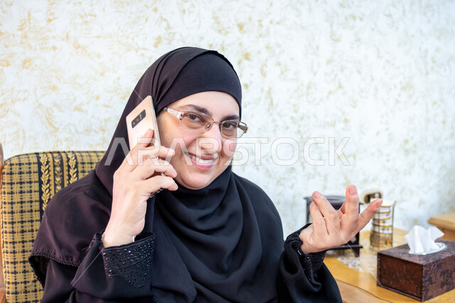 Gestures of joy and happiness upon hearing good news, the use of modern techniques and technology in daily life, a close-up of a Saudi Arabian Gulf woman wearing the traditional hijab and abaya and wearing glasses sitting on a wooden sofa holding a mobile phone and making a phone call