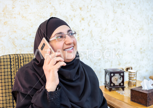 إيماءات السرور والسعادة بسماع أخبار مفرحة، استخدام التقنيات الحديثة والتكنولوجيا في الحياة اليومية،  صورة مقربة لإمرأة عربية خليجية سعودية ترتدي الحجاب والعباءة التقليدية وتضع النظارة الطبية تجلس على أريكة خشبية وتمسك الهاتف المحمول وتجري مكالمة هاتفية