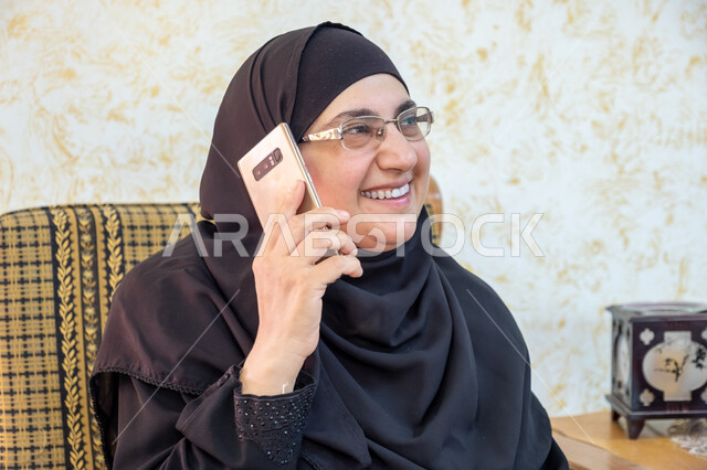 إيماءات السرور والسعادة بسماع أخبار مفرحة، استخدام التقنيات الحديثة والتكنولوجيا في الحياة اليومية،  صورة مقربة لإمرأة عربية خليجية سعودية ترتدي الحجاب والعباءة التقليدية وتضع النظارة الطبية تجلس على أريكة خشبية وتمسك الهاتف المحمول وتجري مكالمة هاتفية