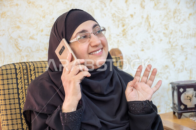 إيماءات السرور والسعادة بسماع أخبار مفرحة، استخدام التقنيات الحديثة والتكنولوجيا في الحياة اليومية،  صورة مقربة لإمرأة عربية خليجية سعودية ترتدي الحجاب والعباءة التقليدية وتضع النظارة الطبية تجلس على أريكة خشبية وتمسك الهاتف المحمول وتجري مكالمة هاتفية