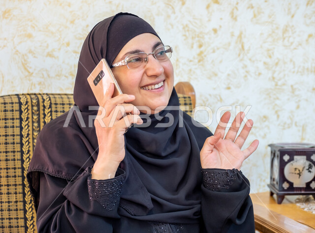 إيماءات السرور والسعادة بسماع أخبار مفرحة، استخدام التقنيات الحديثة والتكنولوجيا في الحياة اليومية،  صورة مقربة لإمرأة عربية خليجية سعودية ترتدي الحجاب والعباءة التقليدية وتضع النظارة الطبية تجلس على أريكة خشبية وتمسك الهاتف المحمول وتجري مكالمة هاتفية