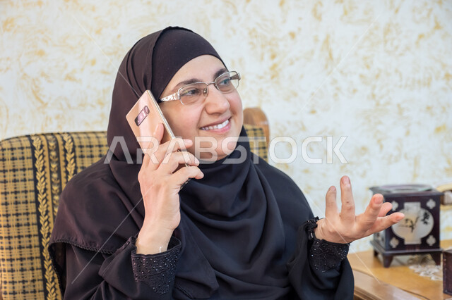 إيماءات السرور والسعادة بسماع أخبار مفرحة، استخدام التقنيات الحديثة والتكنولوجيا في الحياة اليومية،  صورة مقربة لإمرأة عربية خليجية سعودية ترتدي الحجاب والعباءة التقليدية وتضع النظارة الطبية تجلس على أريكة خشبية وتمسك الهاتف المحمول وتجري مكالمة هاتفية