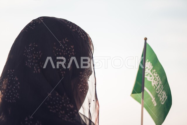 حب الوطن الحفاظ على هوية البلاد، صورة من الخلف لامرأة عربية خليجية سعودية محجبة ترتدي العباءة وتحمل علم السعودية، اليوم الوطني لتوحيد السعودية، العيد الوطني السعودي 8 ربيع الاول، يوم العلم السعودي 11 مارس، خلفية بيضاء