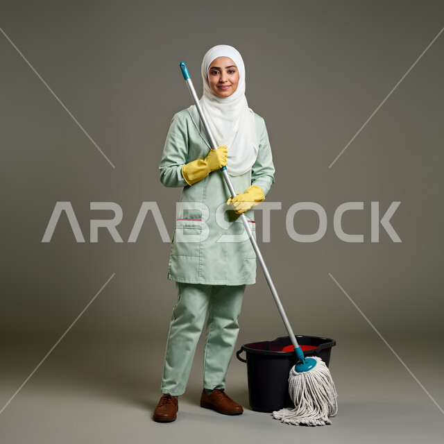 المهام والأعمال المنزلية الروتينية, بورتريه لعاملة نظافة محجبة ترتدي الزي الخاص بالعمل والقفازات تقوم بمسح الأرضية، مهن و وظائف نسائية, النظر إلى الكاميرا بإيماءات السعادة و السرور, الجسم بالطول الكامل, خلفية رمادية