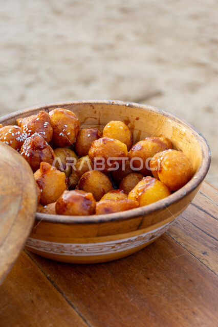 كرات العجين المقلية الصغيرة المرشوشة بالسمسم، الحلوى الشهية واللذيذة، وصفات وأطباق الحلويات والسكريات، ضيافة المناسبات والأعياد العربية، طبق خشبي من اللقيمات ذهبية اللون، حلا الزلابيات الشرقي المغموسة بدبس التمر