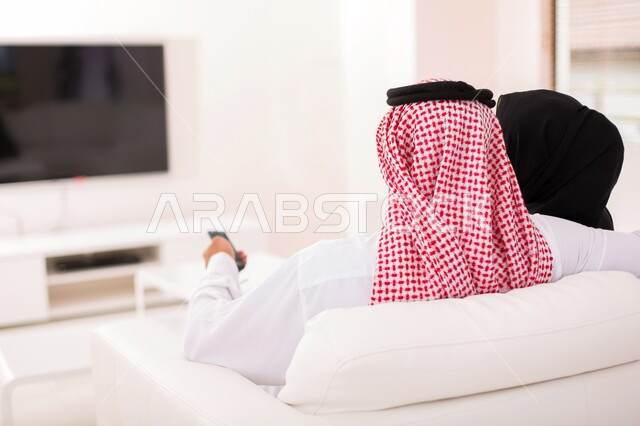 مفهوم الترابط و التفاهم الأسري، صورة مقربة من الخلف لزوجان عربيان خلجيان سعوديان يجلسان على الاريكة يقومان بمشاهدة التلفاز ،متابعة الافلام والمسلسلات، استخدام التكنولوجيا بالترفيه، ايماءات الراحة والاسترخاء، أجواء اسرية سعيدة في المنزل