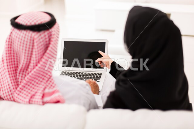 مفهوم الترابط و التفاهم الأسري، صورة مقربة من الخلف لزوجان عربيان خلجيان سعوديان يجلسان على الاريكة يقومان بمشاهدة التلفاز ،متابعة الافلام والمسلسلات، استخدام التكنولوجيا بالترفيه، ايماءات الراحة والاسترخاء، أجواء اسرية سعيدة في المنزل