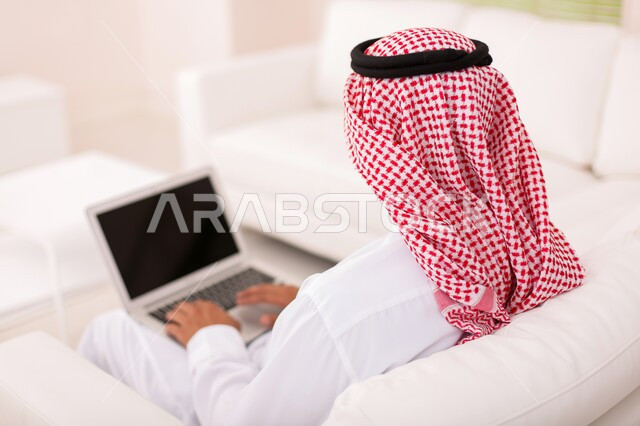 The concept of remote work, a close-up photo from the back of a Saudi Gulf Arab man wearing a shemagh and traditional thobe, completing tasks via a laptop, using modern devices and technologies in economics and financial matters.