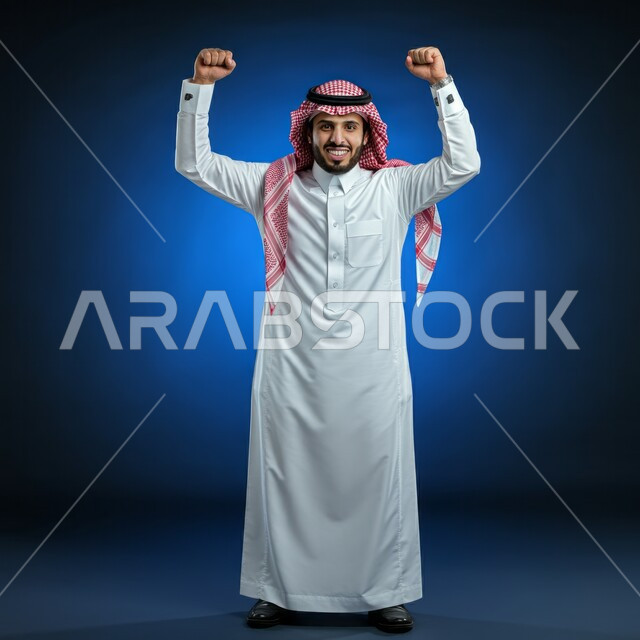مفهوم الانجاز والقوة، المنافسة والتحدي والتشجيع، إيماءات الفرح لسماع خبر ما, النظر الى الكاميرا بتعابير الفوز و تحقيق الأهداف, بورتريه لشاب عربي خليجي سعودي يرتدي الثوب التقليدي والشماغ  يرفع يديه بحركات الحماس و السعادة, صورة الجسم بالطول الكامل, خلفية زرقاء