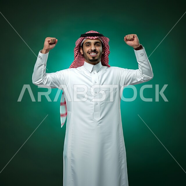 مفهوم الانجاز والقوة، المنافسة والتحدي والتشجيع، إيماءات الفرح لسماع خبر ما, النظر الى الكاميرا بتعابير الفوز و تحقيق الأهداف, بورتريه لشاب عربي خليجي سعودي يرتدي الثوب التقليدي والشماغ  يرفع يديه بحركات الحماس و السعادة, خلفية خضراء