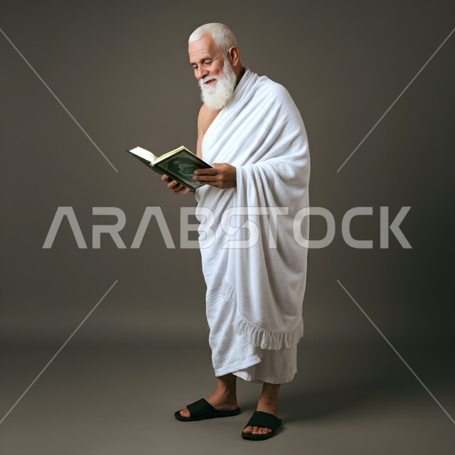 العبادة والتقرب من الله تعالى، بورتريه لرجل مسن ذو لحية بيضاء يرتدي ملابس الإحرام ، الوقوف بابتسامة دافئة وقراءة القران من المصحف الشريف، أداء مناسك الحج والعمرة، الركن الخامس من أركان الاسلام، الطول الكامل للجسم، خلفية رمادية