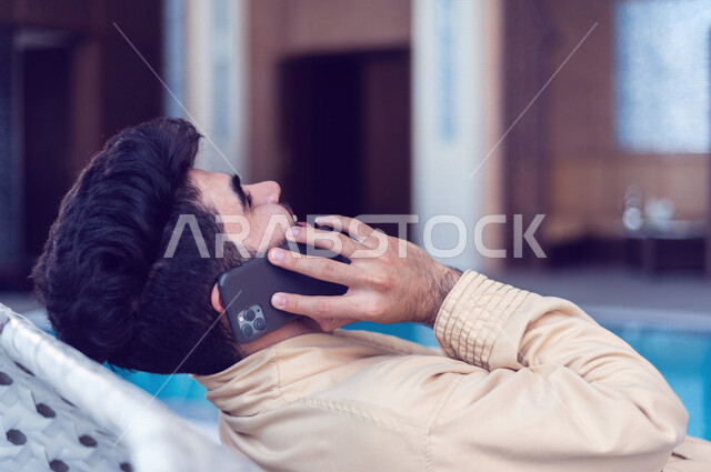 شاب عربي سعودي، الجلوس والاسترخاء في المسبح، اجراء اتصال من خلال الهاتف المحمول