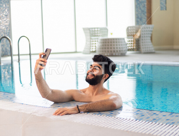 شاب عربي سعودي، متعة السباحة، التدرب على السباحة، الجلوس والاسترخاء في المسبح، التقاط صورة سلفي من خلال الهاتف المحمول