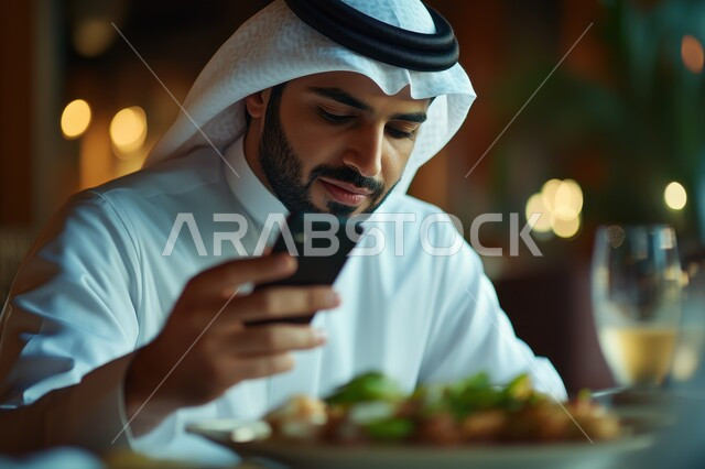 العزائم والولائم في الأعياد والمناسبات الإسلامية، المقبلات والأطباق والأصناف العربية الشرقية اللذيذة، صورة مقربة لرجل عربي خليجي سعودي يرتدي الثوب التقليدي والغترة يجلس على مائدة الطعام ويقوم بالتقاط صورة لطبقه عبر الهاتف المحمول، أجواء شهر رمضان المبارك