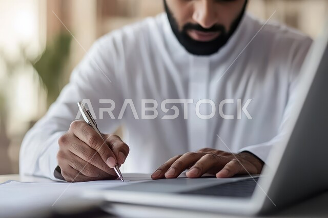 Integrating work with technology and technology, administrative office jobs and professions, managing and organizing business affairs through technical devices, close-up image of a Saudi Arabian Gulf businessman wearing traditional clothing writing down work achievements and next to him the laptop
