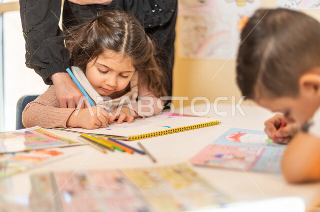 A close-up picture of a Saudi Arabian Gulf boy and girl who is creative in the activity of coloring drawings with the help of the teacher, artistic and recreational activities, education and creativity, skills development, nursery school, kindergarten.