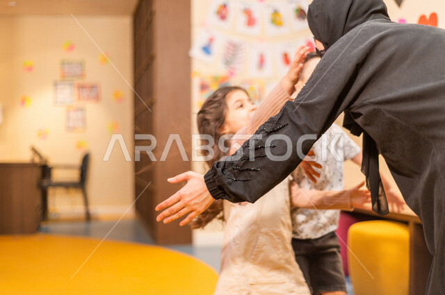 صورة مقربة لمعلمة عربية خليجية سعودية تستقبل بحفاوة طفلان عربيان خليجيان سعوديان ، اتباع اجراءات الوقاية و السلامة ، حب و فرح ، آمان و طمأنينة ، حضانة أطفال ، روضة أطفال .