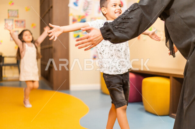 صورة مقربة لمعلمة عربية خليجية سعودية تستقبل بحفاوة طفلان عربيان خليجيان سعوديان ، حب و فرح ، آمان و طمأنينة ، حضانة أطفال ، روضة أطفال .