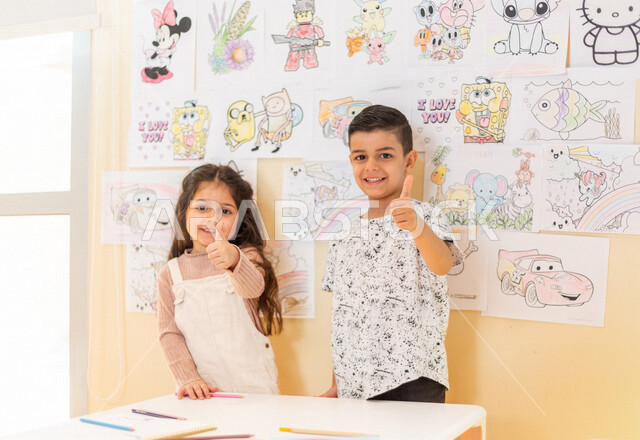 صورة مقربة لطفلان عربيان خليجيان سعوديان يقفان أمام مجموعة من الصور الملونة ، ايماءات و تعابير تدل على السعادة و الانجاز ، أنشطة فنية و ترفيهية ، تعليم و ابداع ، تنمية مهارات ، حضانة أطفال ، روضة أطفال .