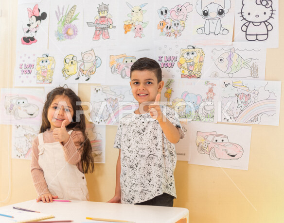 A close-up of two Saudi Arabian Gulf children standing in front of a group of colorful pictures, gestures and expressions indicating happiness and achievement, artistic and recreational activities, education and creativity, skills development, nursery sch