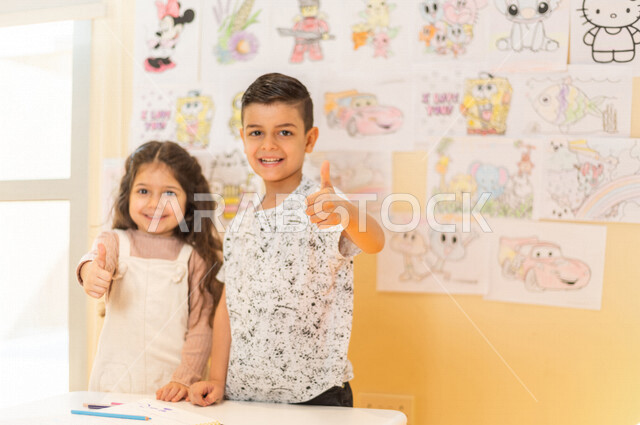 Two Saudi Arabian Gulf children standing in front of a group of colorful pictures, gestures and expressions indicating happiness and achievement, artistic and recreational activities, education and creativity, skills development, nursery school, kindergar