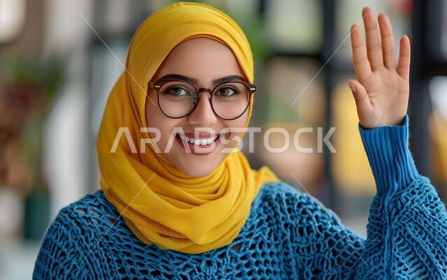 الشعور بالفرح والسرور، مفهوم الأنوثة والاهتمام بالمظهر الخارجي، صورة مقربة لشابة عربية خليجية سعودية محجبة ترفع يدها للاعلى ترتدي النظارة الطبية تنظر الى الكاميرا بإيماءات البهجة والسعادة، المشاركة في النقاشات والحوارات