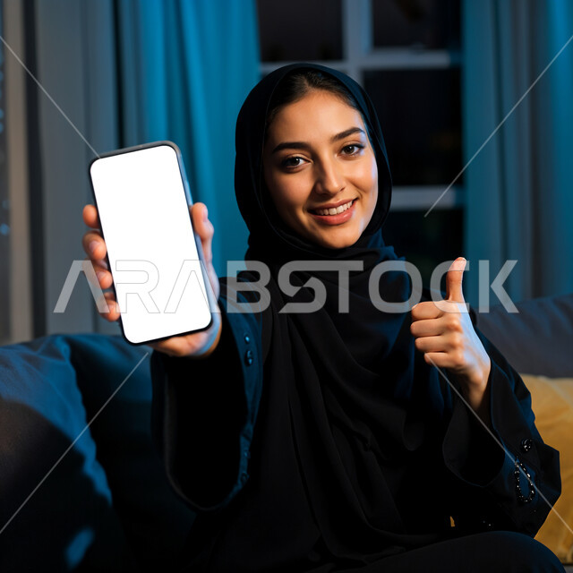 Using modern electronic technologies and devices, promotional marketing ads via mobile, close-up image of a smiling young Saudi Arabian Gulf woman wearing a black abaya and hijab displaying a mobile phone with a blank white screen looking at the camera with gestures of pleasure and happiness