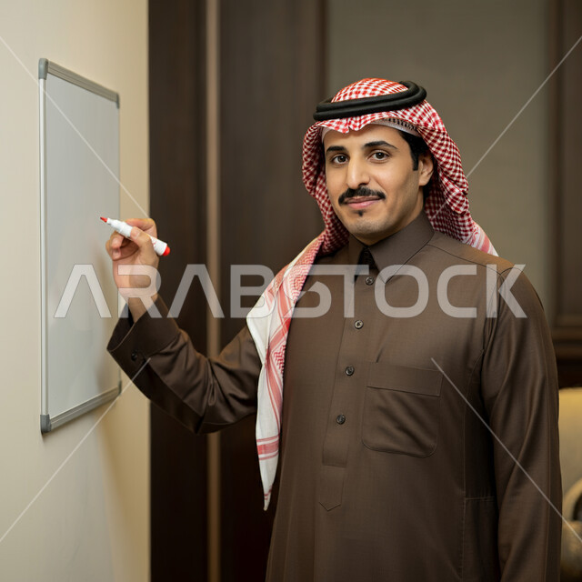 مفهوم التعليم في المملكة العربية السعودية، النظر إلى الكاميرا بإيماءات السعادة و السرور, بورتريه لمعلم صف عربي خليجي سعودي مبتسم يرتدي الثوب التقليدي والشماغ يكتب على لوح أبيض، تدوين الملاحظات وكتابة الأبحاث العلمية