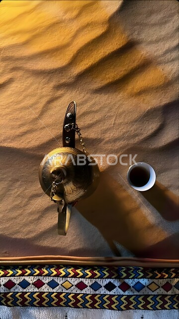 A golden copper Arabic coffee dallah decorated in the middle of the soft golden sand, ancient antiques of ancient style, popular customs and traditions in the Kingdom of Saudi Arabia, warm hospitality and good reception, desert nature background.