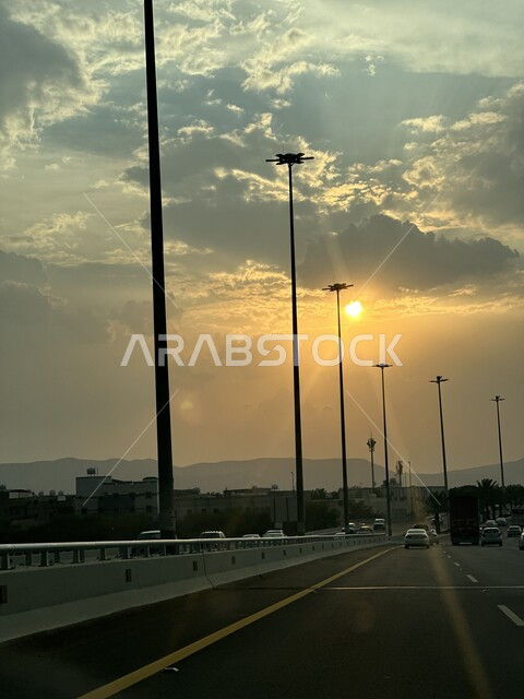 حركة مرور السيارات في الشوارع والطرقات الاسفلتية، منظر شروق الشمس بين الغيوم البيضاء في السماء، أعمدة الإنارة الطرقية، الاهتمام بالتشجير والزراعة في المدينة المنورة في المملكة العربية السعودية