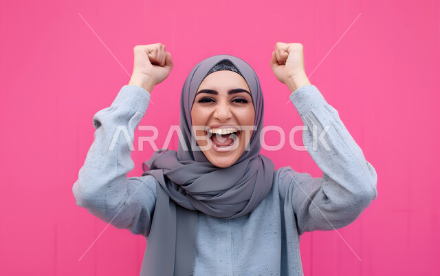 النجاح وتحقيق الأهداف، تعابير السرور والسعادة، بورتريه مقرب لامرأة عربية خليجية سعودية محجبة مبتسمة ترتدي العباءة الملونة تنظر إلى الكاميرا بايماءات وجه ويدين تدل على الفوز والإنتصار، شابة سعودية انيقة، خلفية وردية
