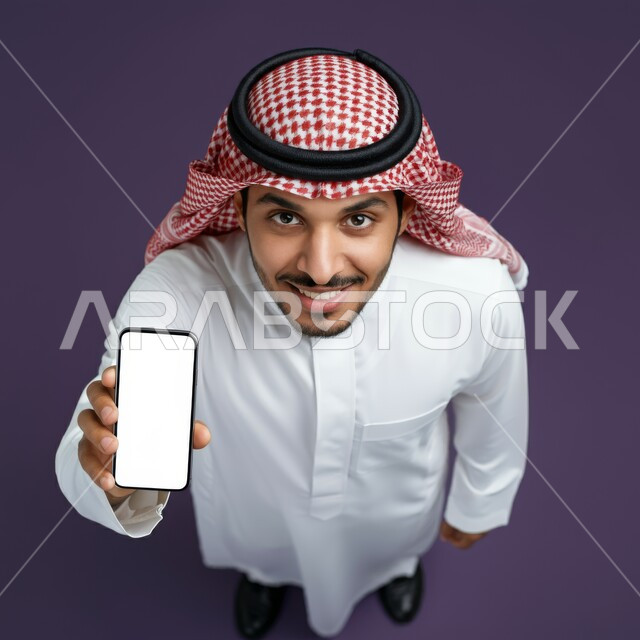 استخدام الأجهزة التقنية الحديثة للتسلية و الترفيه, النظر إلى الكاميرا بإيماءات السعادة و السرور, بورتريه علوي عامودي لشاب عربي خليجي سعودي مبتسم يرتدي الثوب التقليدي و الشماغ يمسك بيده الهاتف المحمول, عرض شاشة بيضاء فارغة عبر الجوال, خلفية إرجوانية