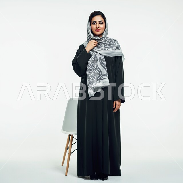 The concept of femininity, elegance and interest in external appearance, portrait of a smiling Saudi Arabian Gulf woman wearing a hijab and an abaya standing in front of a chair looking at the camera, full-length body image, white background