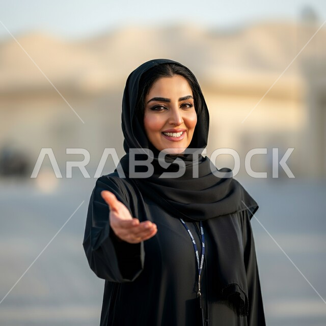 مفهوم الثقة بالنفس, تصاميم أزياء للمحجبات عصرية حديثة، الاهتمام بالمظهر الخارجي و الأناقة، صورة مقربة لامرأة عربية خليجية سعودية مبتسمة محجبة ترتدي عباءة سوداء تنظر الى الكاميرا بايماءات السرور ترفع يدها بحركة تدل على الترحيب والاستقبال