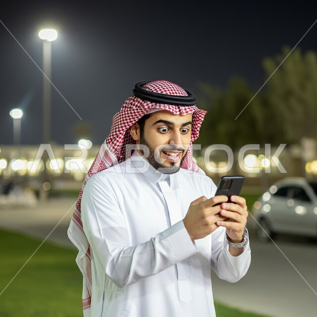 Shocked by hearing sudden news, using modern technical devices, a picture of a smiling Saudi Arabian Gulf young man wearing traditional thobe and shemagh holding a mobile phone in his hand, astonishment and shock at something happening