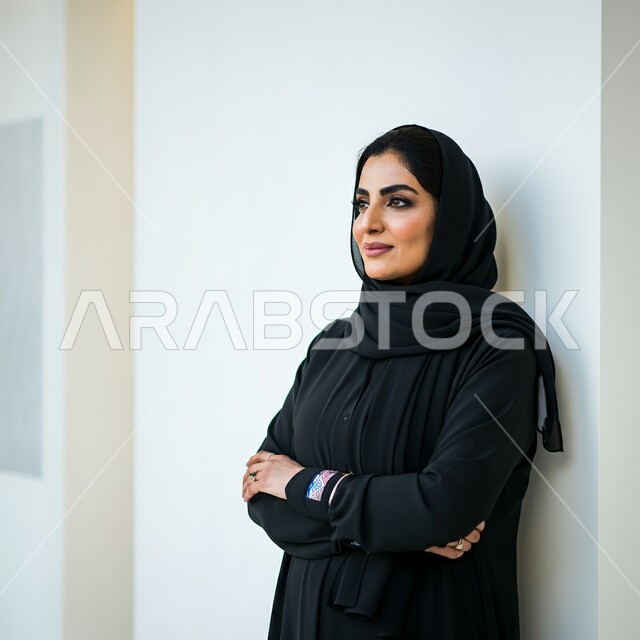 مفهوم الأنوثة والنعومة، الاهتمام بالأناقة والمظهر الخارجي، صورة لامرأة عربية خليجية سعودية مبتسمة محجبة ترتدي العباءة السوداء تنظر الى مكان ما بتعابير السرور والثقة بالنفس، الوقوف بإيماءات أيدي متقاطعة