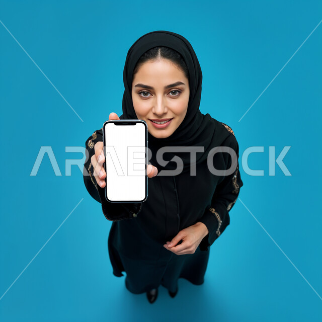 الذكاء الاصطناعي الأجهزة التقنية الحديثة، الاهتمام بالأناقة والمظهر الخارجي، بورتريه مقرب لامرأة عربية خليجية سعودية مبتسمة محجبة ترتدي العباءة السوداء تحمل بيدها الهاتف المحمول و تنظر إلى الكاميرا بإيماءات السعادة و السرور, عرض شاشة بيضاء فارغة عبر الجوال, خلفية زرقاء