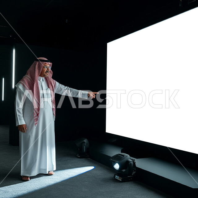 الشرح والتوضيح عبر البروجيكتر، استخدام التقنيه والتكنولوجيا الحديثه في الاجتماعات، مفهوم الإعلان والتسويق، الترويج للخدمات والمنتجات، موك اب مستطيل أبيض، لوح إعلاني فارغ ، صورة لرجل عربي خليجي سعودي يرتدي الثوب التقليدي يشير الى الشاشة