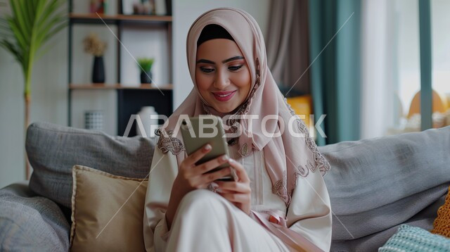  قضاء وقت الفراغ بالتواصل مع الأصدقاء، صورة مقربة لإمرأة عربية خليجية سعودية مبتسمة ترتدي الحجاب تقوم باستعمال الهاتف المحمول، استخدام الأجهزة والتقنيات الحديثة والمتطورة, تصفح المواقع الالكترونية والتواصل الاجتماعي، الجلوس على الكنبة والاستمتاع في وقت الراحة