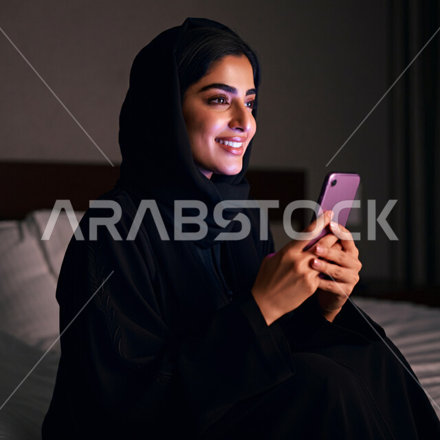 Online shopping through applications, a close-up portrait of a veiled Saudi Arabian Gulf woman, smiling, holding a mobile phone in her hand, spending enjoyable times exchanging news and conversations, communicating and checking on friends, expressions of joy when hearing good news.