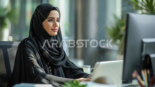 دمج الأجهزة التقنية الحديثة و المتطورة مع الأعمال المكتبية, مهن و وظائف في السعودية, امرأة عربية خليجية سعودية مبتسمة محجبة ترتدي العباءة السوداء تجلس على الطاولة داخل المكتب و تقوم بإنجاز العمل على جهاز الحاسوب