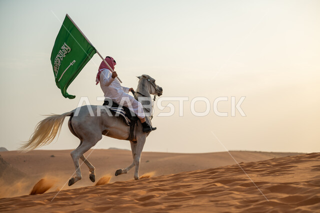 يوم العلم السعودي 11 مارس، علاقة أبناء الوطن بالخيل السعودي الأصيل، رجل عربي خليجي سعودي يرتدي الشماغ والزي التقليدي يتجول على ظهر الحصان يمسك بيده العلم في الصحراء وقت غروب الشمس، الإحتفال بذكرى ال93 لليوم الوطني السعودي 2023، العيد الوطني 23 سبتمبر