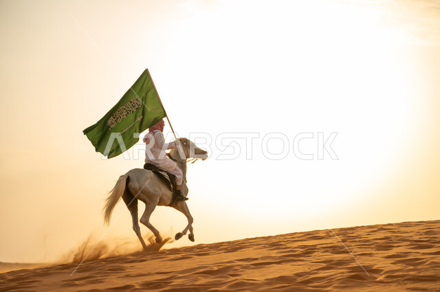 Saudi Flag Day March 11, the nation’s relationship with the purebred Saudi horse, a Saudi Arabian Gulf man wearing a shemagh and traditional costume walking around on the back of a horse holding a flag in the desert at sunset, celebrating the 93rd anniver