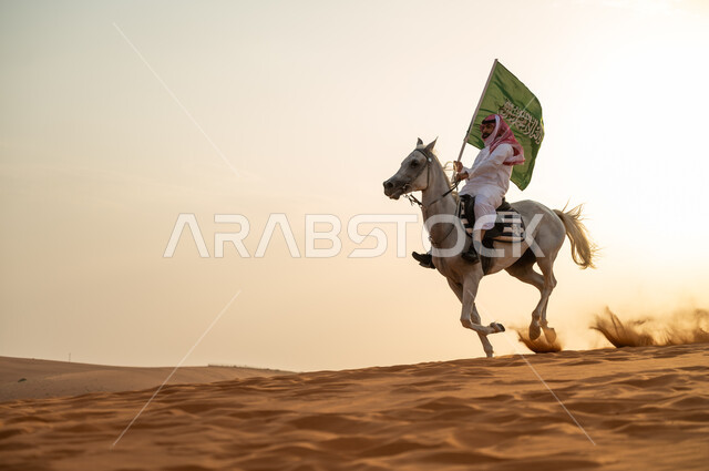 Saudi Flag Day March 11, the nation’s relationship with the purebred Saudi horse, a Saudi Arabian Gulf man wearing a shemagh and traditional costume walking around on the back of a horse holding a flag in the desert at sunset, celebrating the 93rd anniver