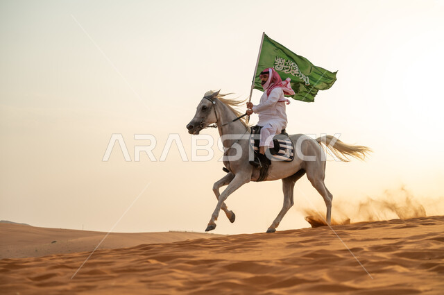 يوم العلم السعودي 11 مارس، علاقة أبناء الوطن بالخيل السعودي الأصيل، رجل عربي خليجي سعودي يرتدي الشماغ والزي التقليدي يتجول على ظهر الحصان يمسك بيده العلم في الصحراء وقت غروب الشمس، الإحتفال بذكرى ال93 لليوم الوطني السعودي 2023، العيد الوطني 23 سبتمبر